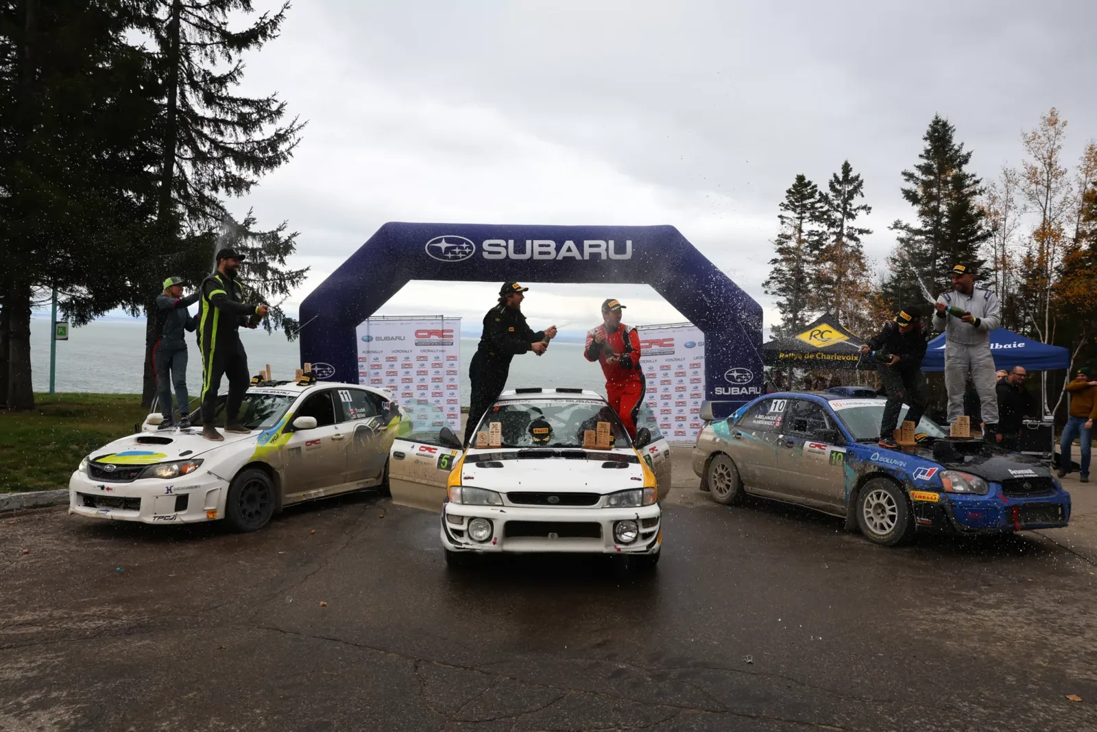 Three crews celebrating on the Rallye de Charlevoix podium, with the St. Lawrence River behind them