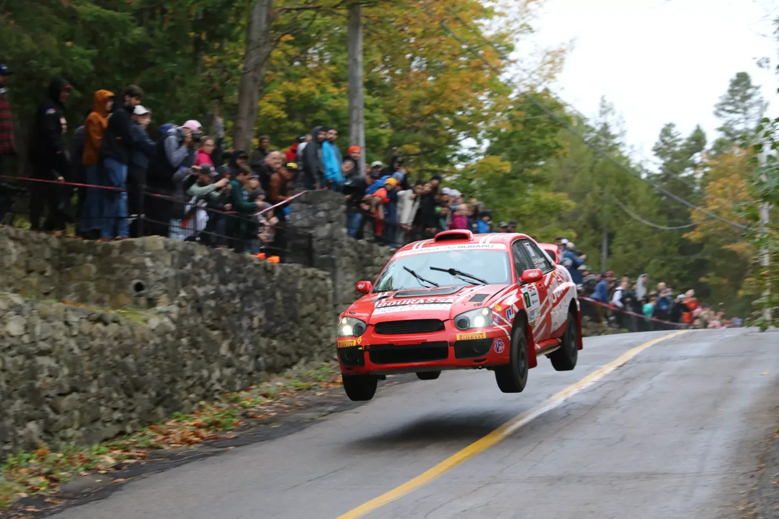Rally car airborne on an asphalt road lined with a stone wall, in front of spectators behind the safety tape