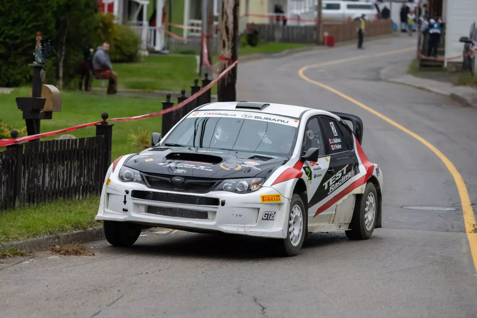 Rally car taking a bend on a village street, between houses and the safety tape